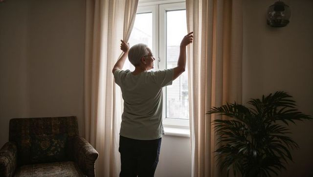 Senior woman opening curtains at bright double-pane window in cozy neutral living room