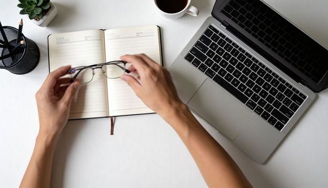 Holding glasses over open lined notebook on minimal desk with laptop, coffee and plant