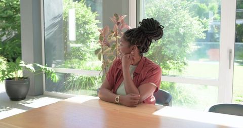 Adult african woman sitting at sunroom table looking out sunlit windows and greenery