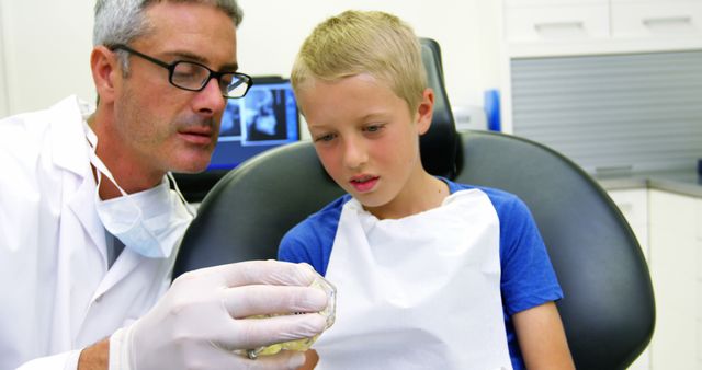 Caucasian male dentist showing dental model to boy patient in chair at dental surgery. Childhood, dentistry, oral care, medical services, hygiene and work, unaltered.