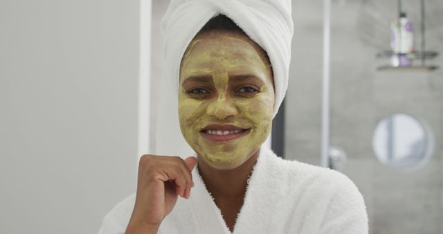 Woman is standing in a spa bathroom wearing a white robe and towel wrap, with a face mask applied. She is smiling and appears relaxed, emphasizing self-care and wellness. This is ideal for promoting skincare products, wellness routines, beauty tutorials, and spa services.