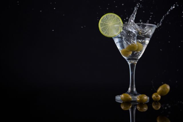 Martini cocktail with olive splashing into the glass and lime garnish on black background. Ideal for use in advertisements, bar menus, cocktail recipes, nightlife promotions, and party invitations.