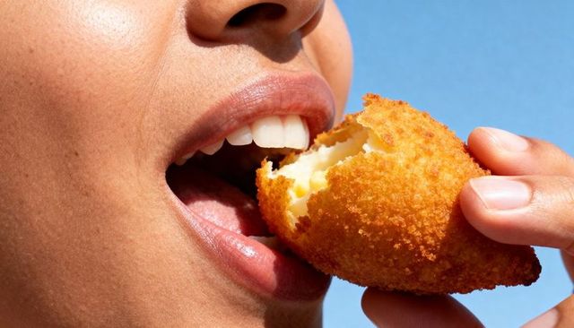 Woman biting golden breadcrumb-coated cheese croquette while holding with fingertips