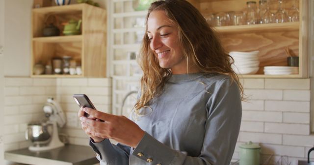 A woman is smiling while using her smartphone in a cozy kitchen. The setting includes warm, wooden shelves and modern appliances, displaying a comfortable and casual environment. This image can be used for themes such as home lifestyle, social media, technology use, communication, and casual domestic moments.