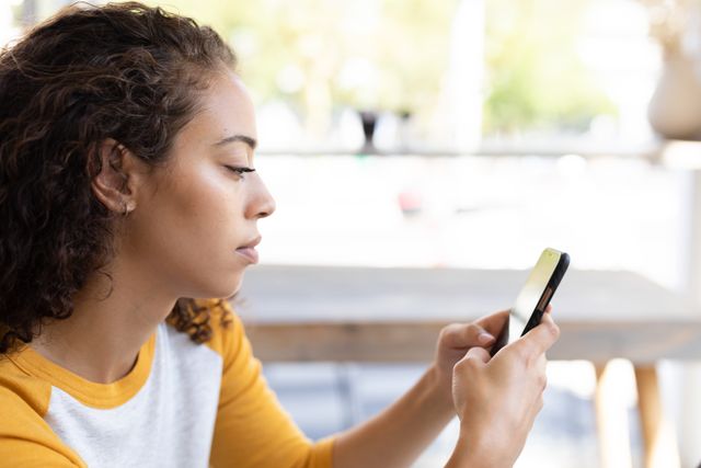 Young woman sitting in a cafe using a smartphone. Ideal for illustrating modern communication, technology use, social media engagement, and casual lifestyle. Suitable for articles, blogs, and advertisements related to technology, connectivity, and cafe culture.