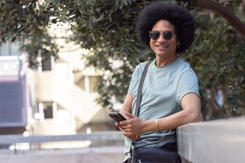 African american man leaning on urban plaza railing smiling while using smartphone