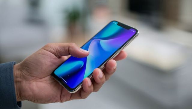 Left hand holding smartphone showing blue purple abstract wallpaper, closeup in home interior