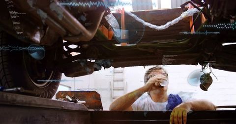 Greasy mechanic drinking from paper cup leaning over inspection pit under lifted car