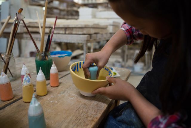 Young girl decorating a ceramic bowl in a pottery workshop. She is using various colors and brushes to create her design. This image can be used for educational materials, art and craft promotions, children's activities, and creative hobby advertisements.