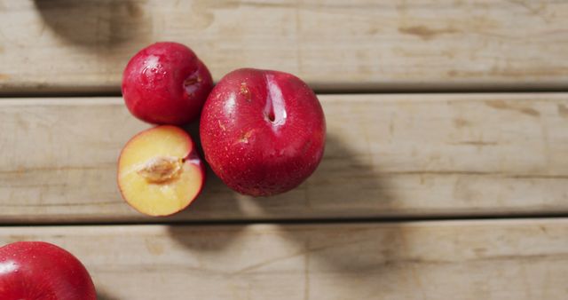 Fresh red plums displayed on a wooden table with one sliced in half, exposing the inner flesh and pit. Suitable for use in advertising healthy foods, nutritional articles, seasonal fruit promotions, or rustic kitchen scenes.