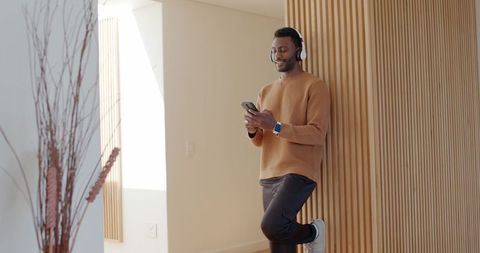 African american man leaning on wooden slat wall checking smartphone wearing headphones