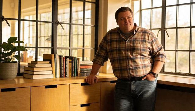 Leaning man wearing plaid shirt and jeans resting on wooden counter in sunlit workspace