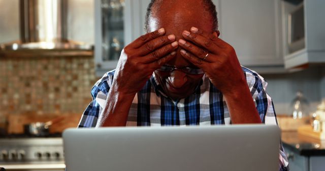 Senior man sitting in modern kitchen, holding his head in frustration while using laptop. He appears stressed and overwhelmed by technology or tasks he is working on. Useful for articles related to elderly and technology, stress management, remote work for seniors, and digital literacy.