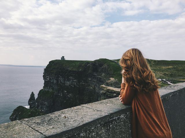 Woman with light brown hair leans on stone wall, overlooking the Cliffs of Moher in Ireland. The cloudy sky creates a peaceful atmosphere. Can be used for travel blogs, brochures, or content highlighting scenic locations and nature experiences.