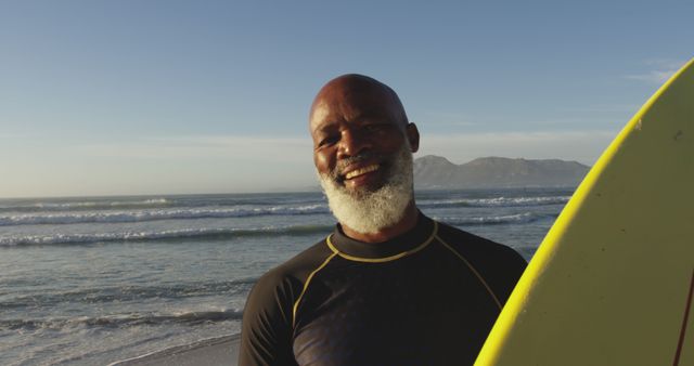Senior man smiling while holding surfboard on beach during sunset. Perfect for promoting active lifestyles for seniors, vacations, seaside activities, and ads targeting mature audiences interested in sports and outdoor hobbies.