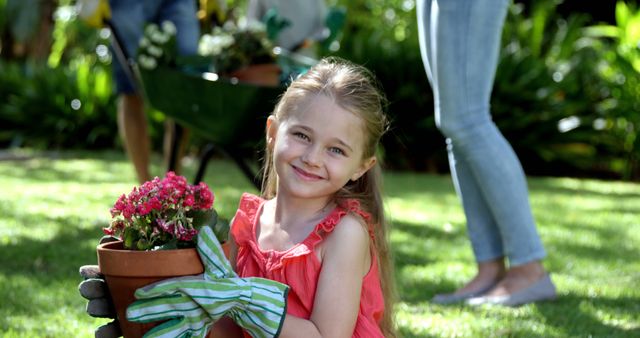 A young Caucasian girl smiles while holding a potted plant in a sunny garden, with copy space. She is learning about gardening, contributing to a family activity outdoors.