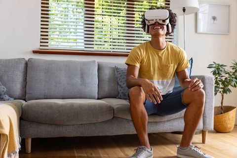 Young man wearing vr headset sitting on sofa enjoying virtual reality in modern home