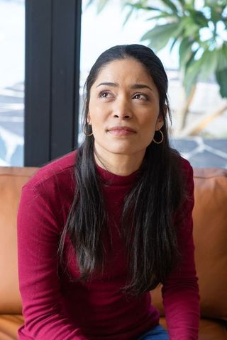 Woman sitting on tan leather sofa looking thoughtful by window wearing burgundy sweater