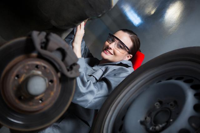 Female mechanic wearing safety glasses smiling while repairing a car wheel in a repair garage. Ideal for use in articles, advertisements, and promotional materials highlighting gender diversity in trades, automotive services, and skilled labor professions.