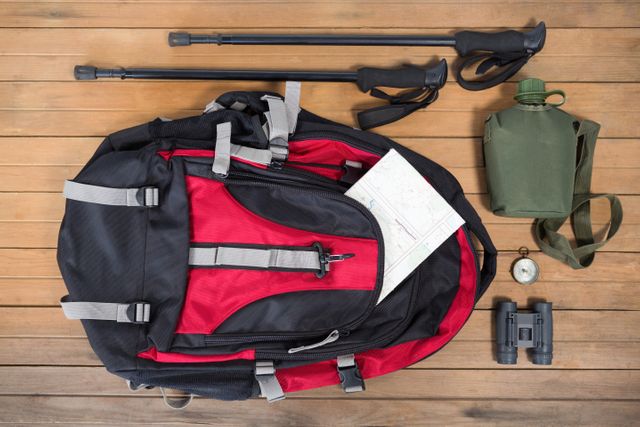 Overhead view of travel gear including a red and black backpack, map, binoculars, compass, canteen, and trekking poles on a wooden floor. Ideal for illustrating travel preparation, outdoor adventures, hiking trips, and camping essentials. Suitable for use in travel blogs, adventure magazines, and outdoor gear advertisements.