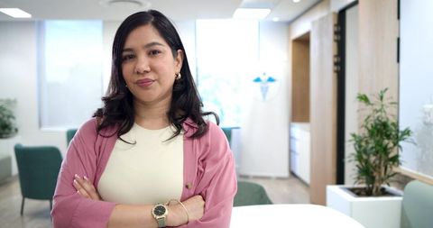 Standing confident woman crossing arms wearing pink cardigan in modern medical clinic