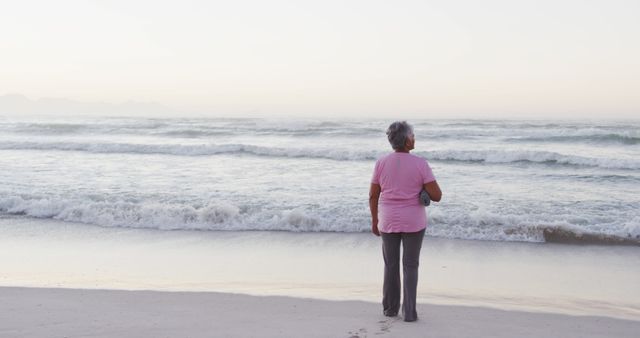 Senior woman in pink shirt standing on beach, gazing at peaceful ocean waves during sunset. Suitable for themes related to retirement, tranquility, leisure, and connection with nature. Ideal for use in travel blogs, wellness articles, and advertisements focusing on peaceful living and relaxation.