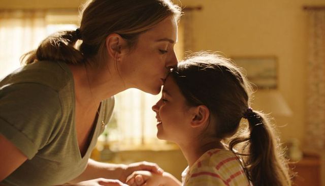 Mother leaning, kissing daughter's forehead in warm backlit cozy home moment of affection