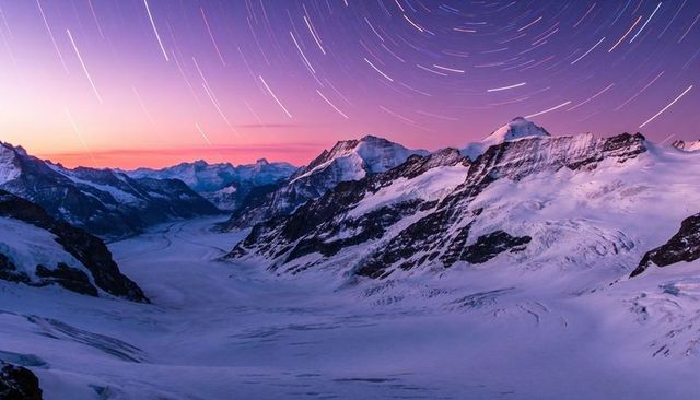 Star trails sweeping over snowcapped alpine glacier valley at twilight purple glow