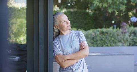 Mature woman leaning on patio post, gazing into sunlit garden, relaxed casual style