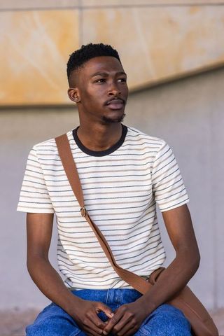 Thoughtful man sitting with crossbody bag wearing striped knit shirt, casual urban portrait