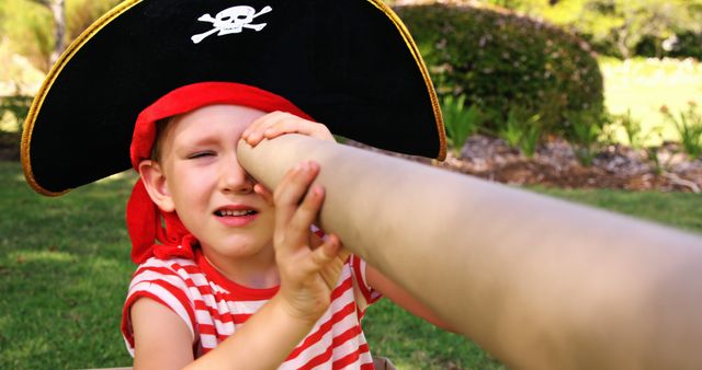A young Caucasian child dressed as a pirate is playfully looking through a hand telescope made by an adult's hand, with copy space. Imaginative play is captured here as the child engages in a fun outdoor activity.