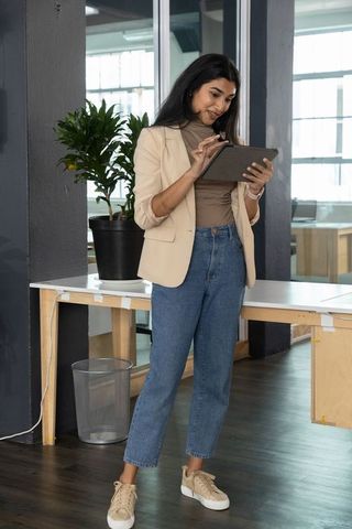 Indian businesswoman tapping tablet standing in modern open-plan office wearing beige blazer