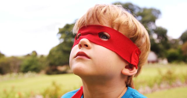 Young boy wearing red superhero mask looking up, standing outdoors. Ideal for concepts involving childhood dreams, playfulness, imagination, and adventure. Perfect for advertising or editorial content related to kids, superhero themes, outdoor activities, summer fun, and children's education.