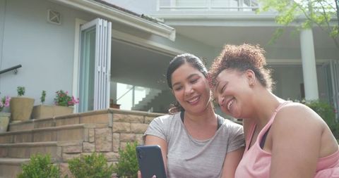 Diverse friends sharing smartphone and smiling on stone steps outside modern home