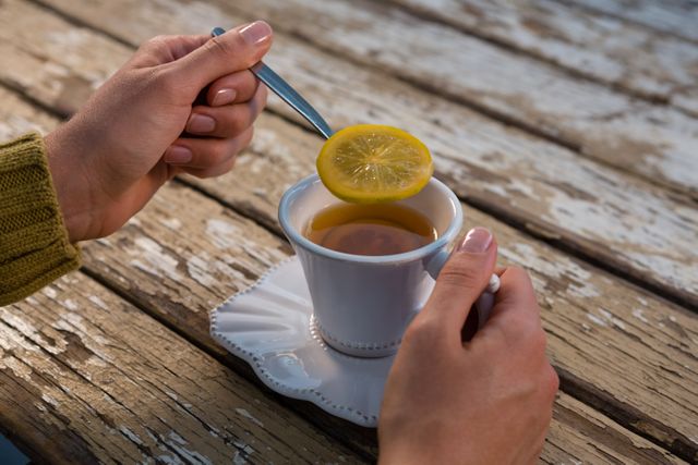 Woman's hands holding a lemon slice over a cup of tea on a rustic wooden table. Ideal for use in lifestyle blogs, wellness articles, and food and beverage promotions. Perfect for illustrating themes of relaxation, morning routines, and cozy moments.