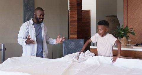 African American father and son making bed together spreading white sheet and smiling