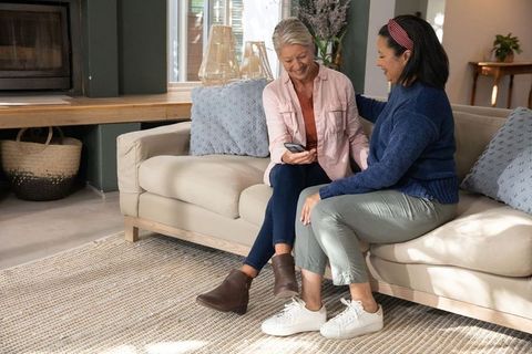 Mature women sharing smartphone on cozy living room sofa, smiling and connecting
