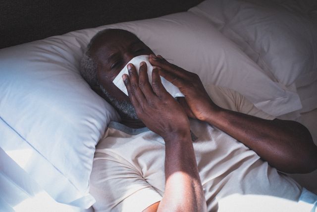 African american senior man lying in bed ill covering face with handkerchief. staying at home in isolation during quarantine lockdown.