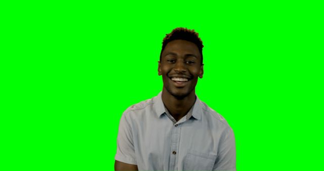 A cheerful young man smiling while standing in front of a green screen, wearing a light-colored casual shirt. Perfect for projects requiring chroma key background, video production, digital content creation, or any related creative purposes where subject isolation and background replacement are needed.