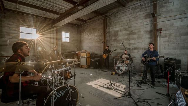 Five-piece rock band rehearsing in industrial warehouse with sun flare and vintage amps