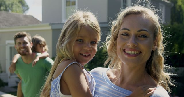 Mom and dad smiling with young children outside home. Mom holding daughter while dad gives piggyback ride to other child. Perfect for advertisements showcasing family life, housing communities, summer activities, parenting articles, and homeownership.