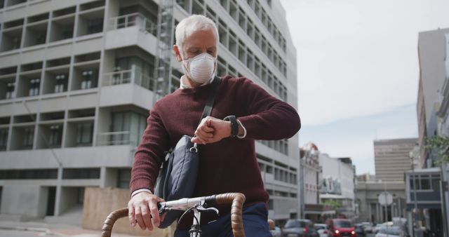 A senior man rides a bicycle in an urban environment while wearing a protective mask. He checks the time on his wristwatch, possibly to stay on schedule. This image can be used in contexts related to urban lifestyles, health and safety, senior fitness, time management, or pandemic-related precautionary measures.