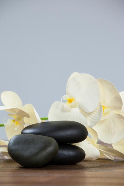 Stacked pebble stones with white orchids on a wooden table create a serene and tranquil scene. Ideal for use in spa and wellness promotions, meditation and relaxation content, and nature-inspired designs. Perfect for conveying themes of balance, harmony, and natural beauty.