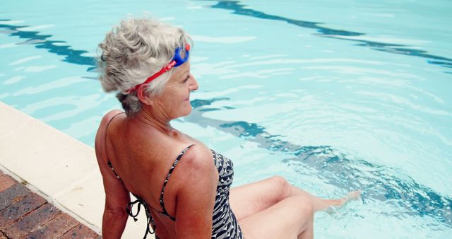 A senior Caucasian woman wearing swimming goggles and a swimsuit sits by the poolside, with copy space. Her relaxed posture and swim attire suggest she is enjoying a leisurely day at the swimming pool.