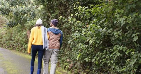 Diverse couple walking arm in arm on mossy trail wearing knit hats and rain boots