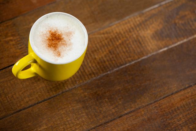 Bright yellow coffee cup filled with frothy cappuccino or latte topped with a sprinkle of cinnamon on rustic wooden table. Perfect for coffee shop promotions, food blogs, cozy morning routines, or beverage advertisements.