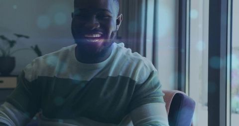 Smiling man sitting in armchair by window with lens flare, moody bokeh, casual sweater
