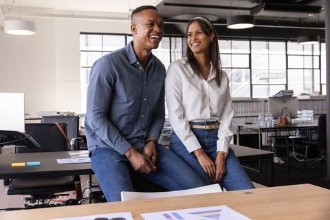 Smiling diverse coworkers sitting on desk in modern open-plan office workspace collaborating