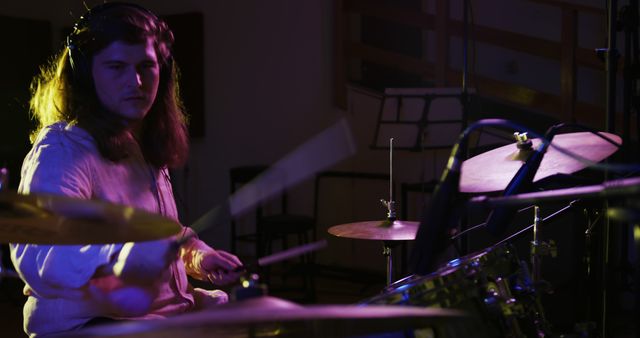 Side view of a Caucasian man with long dark hair wearing headphones and playing a drum kit during a session at a recording studio. Musicians working on producing a song