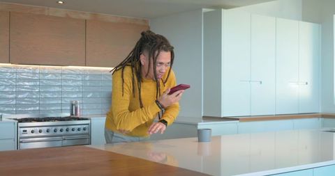 African american man speaking into smartphone while leaning over modern kitchen island